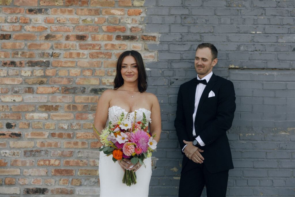 Bride stands holding a colorful wedding bouquet against a brick wall while the groom waits behind her, highlighting bold floral choices and modern wedding colors 2026.