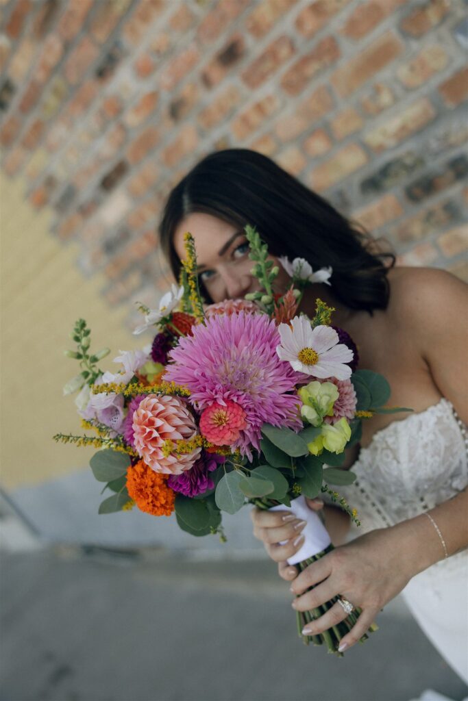 Close-up of a colorful wedding bouquet featuring pink dahlias, orange marigolds, white cosmos, and greenery, arranged with a loose, natural feel.