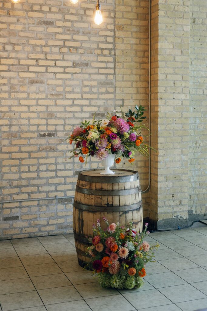 A lush floral arrangement in pinks, oranges, and reds sits in a white urn atop a wooden barrel against an exposed brick wall, a playful nod to wedding colors 2026 that feel warm and expressive.