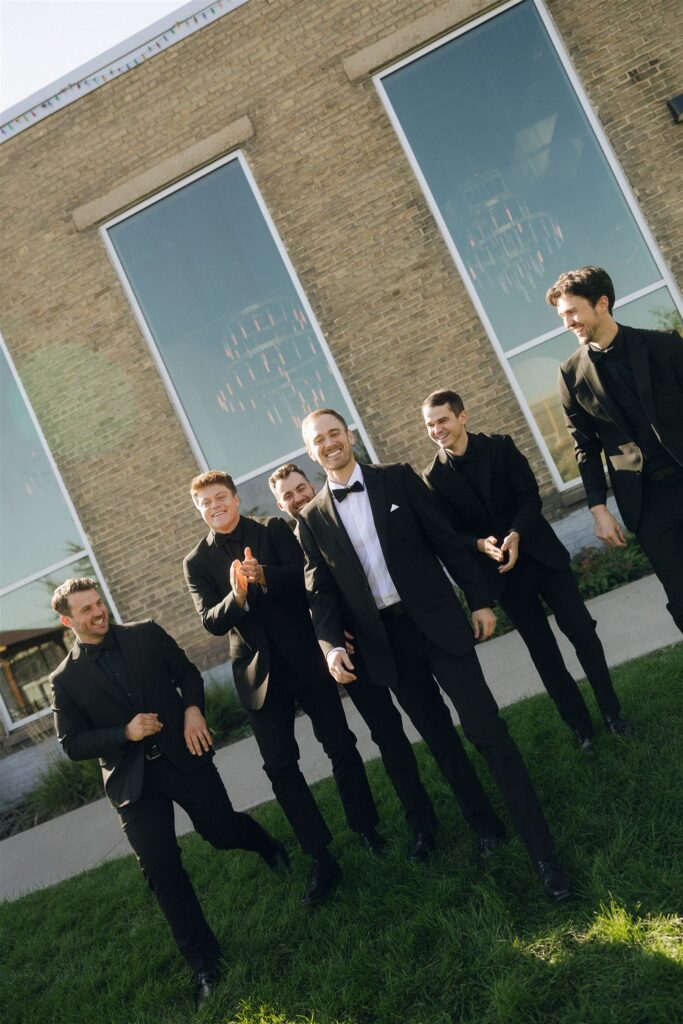 Group of groomsmen in black suits walk together outside a brick venue, laughing and mid-conversation in a relaxed pre-ceremony moment.