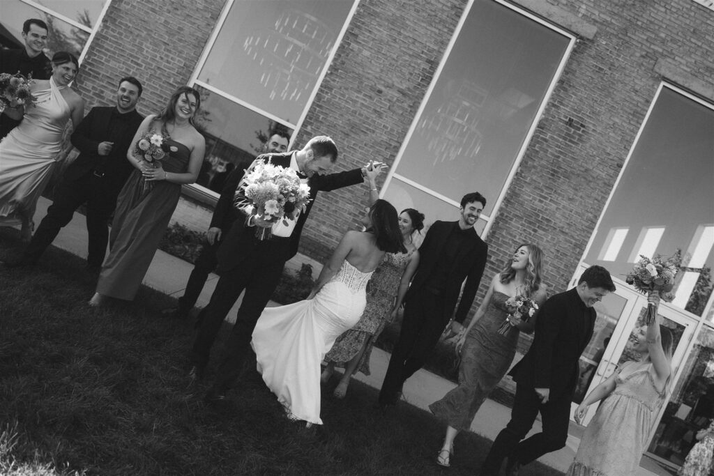 Black-and-white photo of the wedding party walking together outdoors, bouquets and dresses in motion as they laugh and celebrate.
