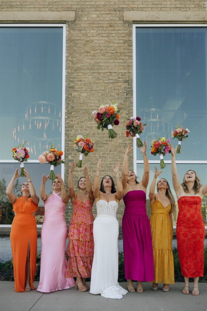 Bride and bridesmaids toss their bouquets into the air in front of a brick building, showcasing playful styling and vibrant wedding colors 2026.