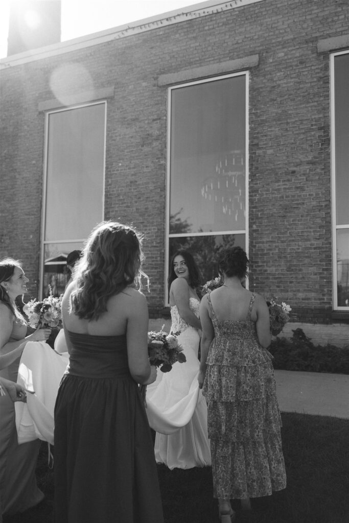 Black-and-white photo of the bride walking outdoors with her bridesmaids, smiling back over her shoulder as dresses and bouquets move in the afternoon light.
