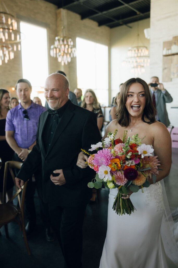 Bride walks down the aisle arm in arm with her father inside an industrial brick venue, holding a colorful bouquet as guests look on.