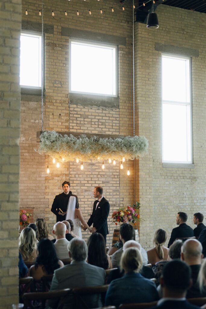 Wide view of an indoor wedding ceremony in a brick venue with hanging greenery and warm string lights, a popular layout featured in wedding planning resources.