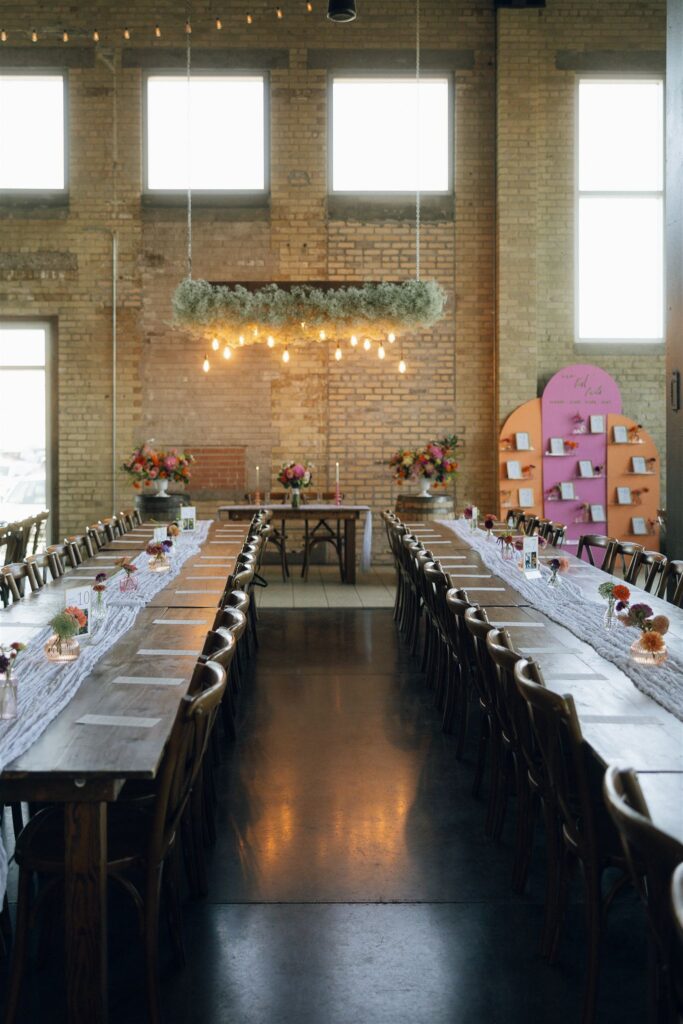 Long wooden reception tables set with simple runners, bud vases, and candles inside a brick-walled venue, styled with intention using thoughtful wedding planning resources.