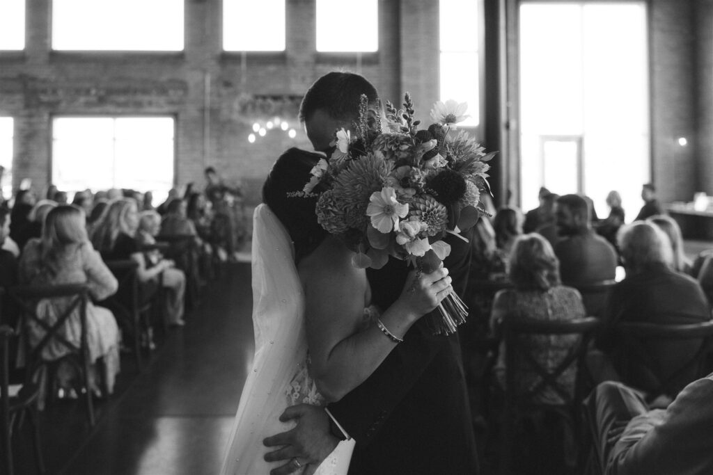 Black-and-white image of the couple embracing during the ceremony, the bride holding her bouquet as guests watch from their seats.