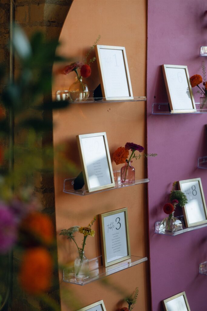 Escort card display with framed table numbers arranged on colorful shelves, accented with small floral arrangements and playful wedding colors 2026.