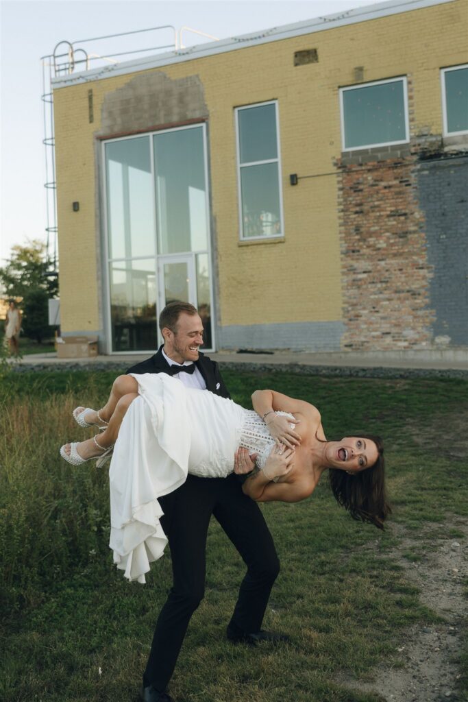 Groom carries the bride across the grass outside an industrial venue, both laughing openly in an unscripted moment between events.
