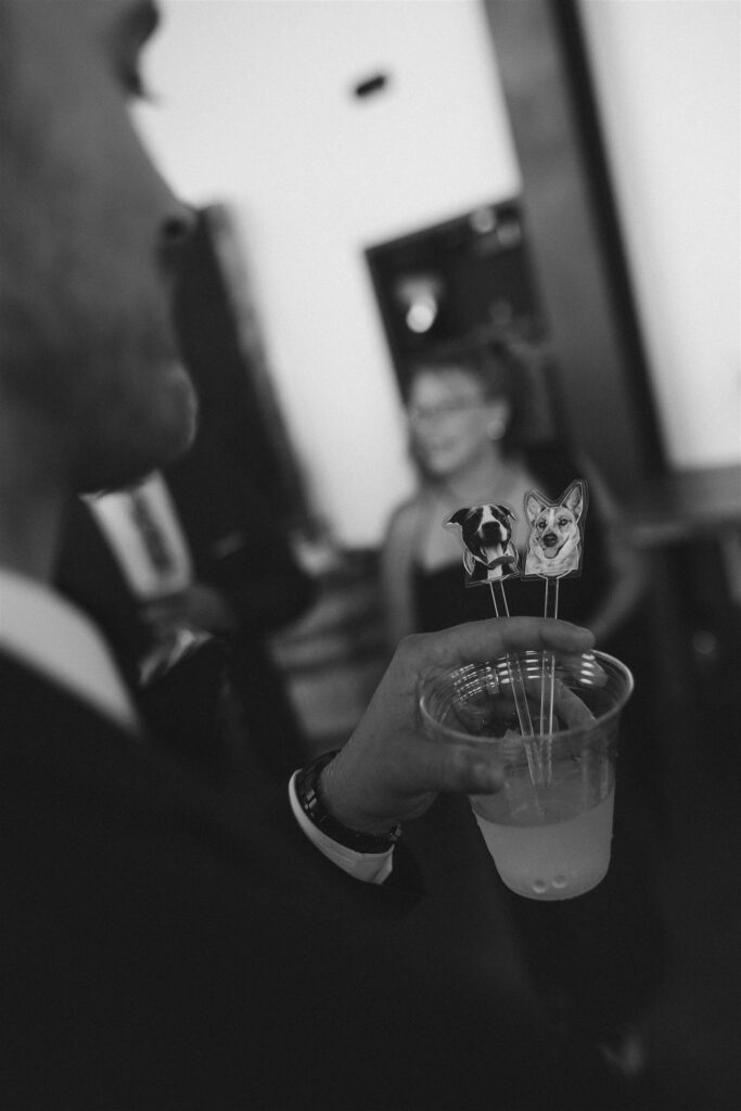 Black and white photo of a groom holding a cocktail with custom dog stir sticks during a wedding reception, showcasing personal details featured in modern wedding planning resources.