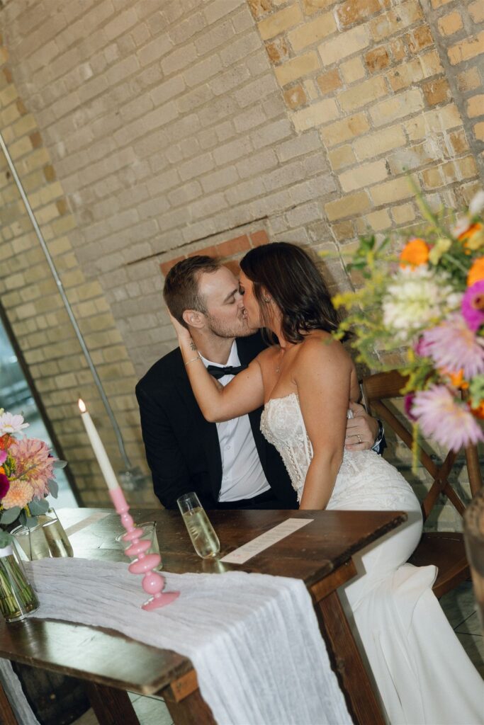 Bride and groom seated at a reception table sharing a kiss beside a colorful wedding bouquet and modern table décor inspired by wedding colors 2026.