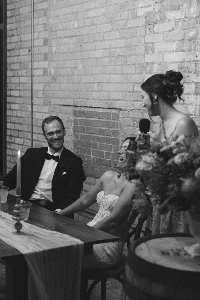 Black and white photo of guests giving heartfelt speeches at a long wooden reception table, emphasizing intimate wedding design and planning resources.