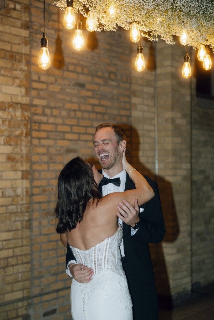 Bride and groom dancing together beneath hanging lights and greenery, surrounded by warm tones and a colorful wedding bouquet aesthetic.
