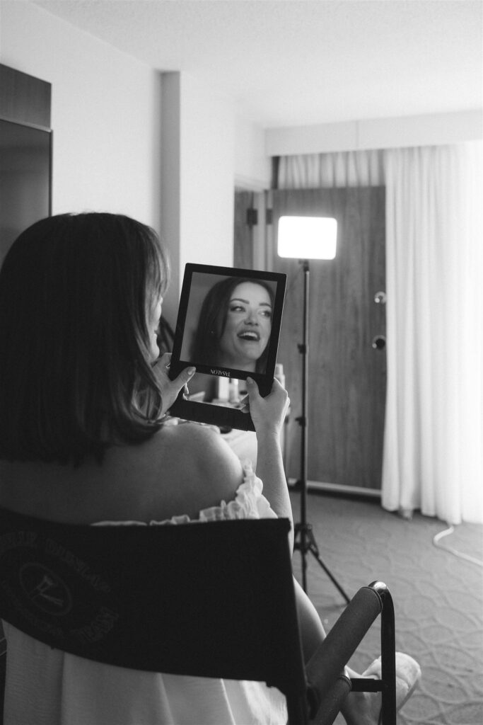 Bride sits in a getting-ready chair holding a handheld mirror, smiling at her reflection during a quiet pre-ceremony moment.