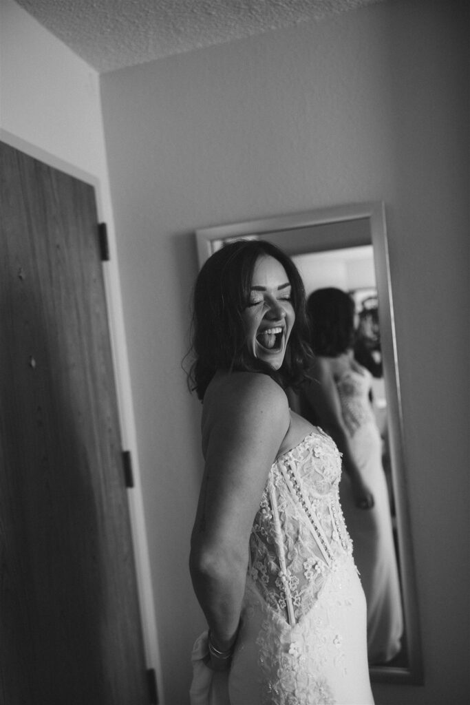 Black-and-white portrait of the bride laughing in front of a full-length mirror, dress partially fastened, capturing joy before the ceremony begins.