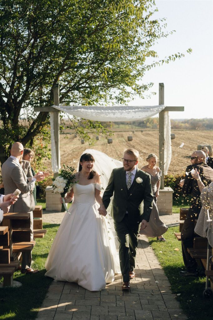 The bride and groom walk hand in hand back down the ceremony aisle at Legacy Hill Farm, smiling as guests toss dried flower petals into the air. A simple wooden ceremony arbor stands behind them, with open farmland stretching into the distance beyond the venue.