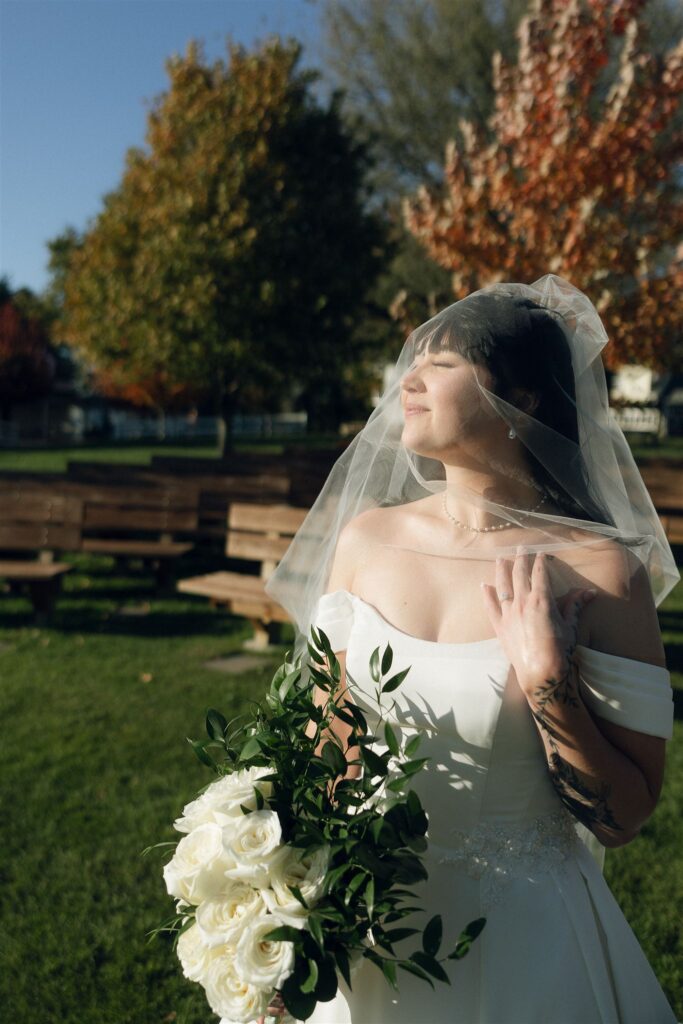 A close portrait of the bride at Legacy Hill Farm, standing in sunlight with her eyes closed. She wears a white off-the-shoulder gown and veil, holding a bouquet of white roses and greenery, with wooden ceremony benches and green grass softly blurred in the background.