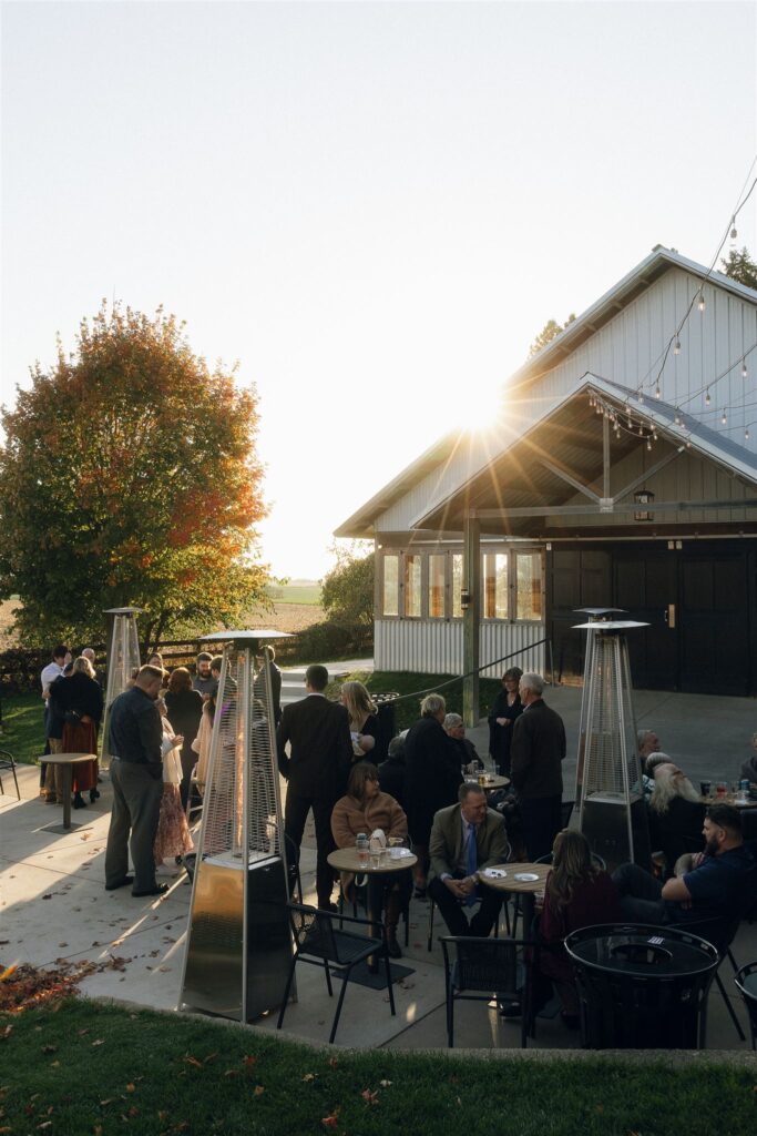 An outdoor cocktail hour scene at Legacy Hill Farm, where guests gather around small round tables on a concrete patio. Tall patio heaters stand among the crowd, and the barn building is visible behind them as the sun sets over the surrounding farmland.