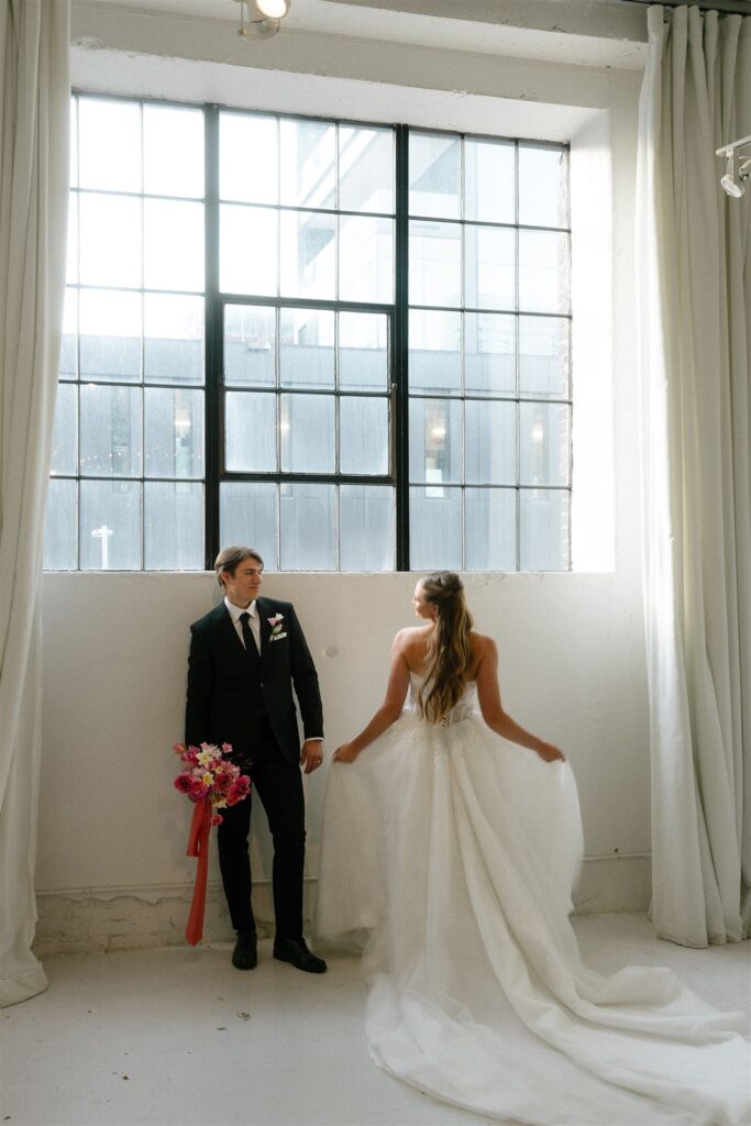 Bride and groom standing beneath tall industrial grid windows at Paris Dining Club, a modern Minnesota wedding venue, with the bride lifting the skirt of her flowing white gown and the groom holding a colorful pink and coral bouquet inside the minimalist white space.