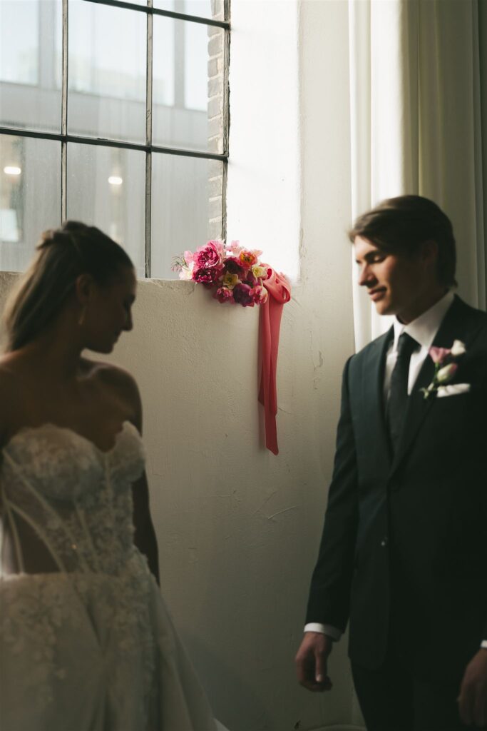 Close-up of a vibrant pink, coral, and yellow wedding bouquet with trailing silk ribbon resting on a windowsill between the bride and groom at Paris Dining Club, showcasing colorful floral design inside one of the best wedding venues in Minnesota.