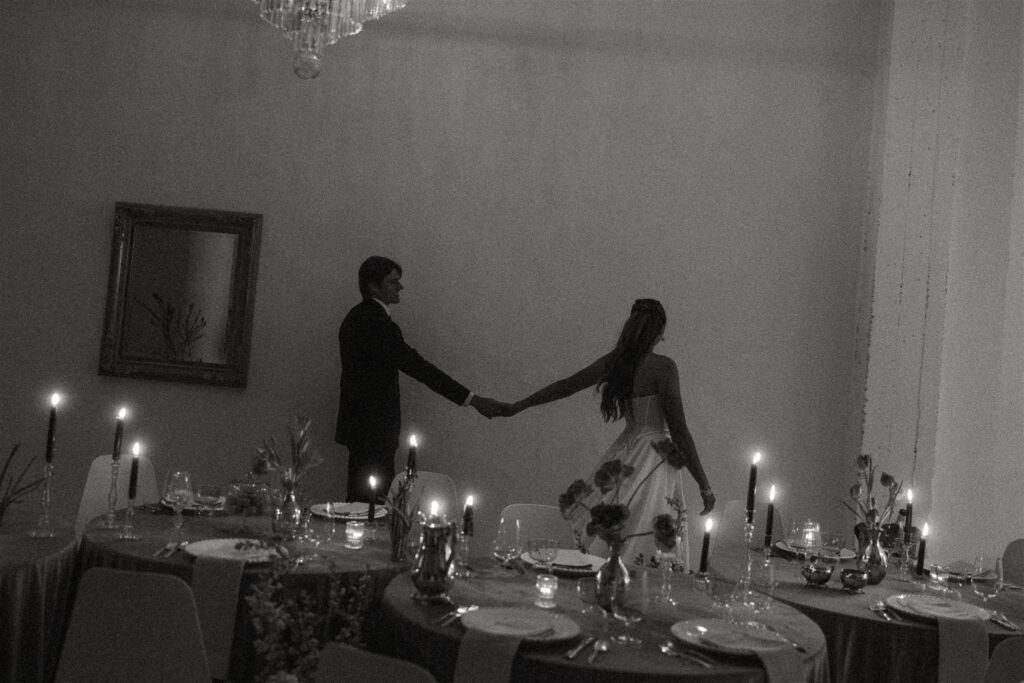 Black-and-white photograph of the bride and groom holding hands and walking between candlelit reception tables at Paris Dining Club, highlighting the moody, editorial atmosphere of this Minnesota wedding venue.