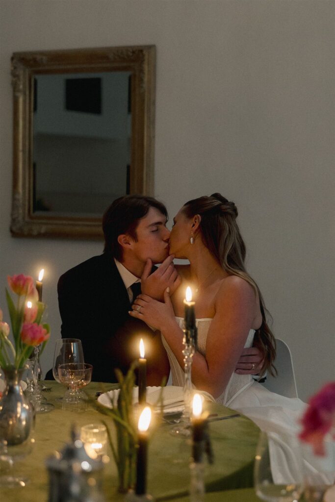 Bride and groom seated together at a candlelit reception table at Paris Dining Club, sharing a kiss surrounded by green velvet table linens, pink florals, glassware, and glowing taper candles at one of the best wedding venues in Minnesota.