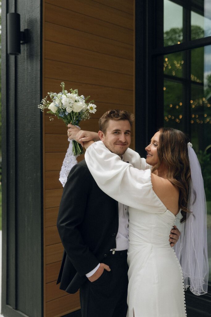 Bride hugging the groom while holding a white floral bouquet outside Woods and Co Weddings and Events, a candid portrait at one of the most scenic Minnesota wedding venues.