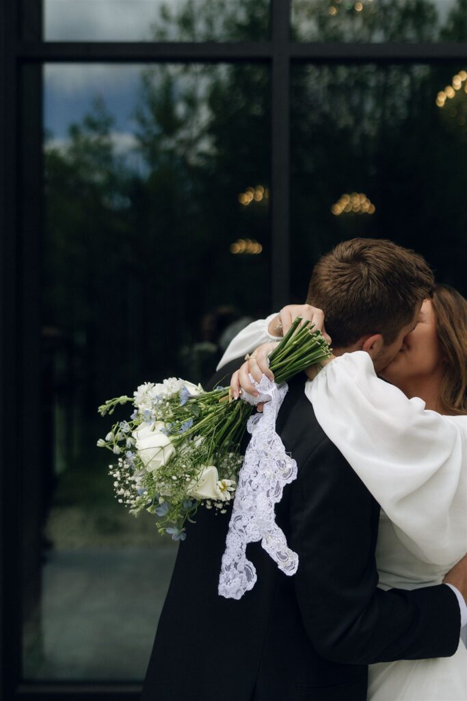 Bride and groom kissing closely, the bride holding a simple white and greenery bouquet tied with lace, captured at Woods and Co Weddings and Events.