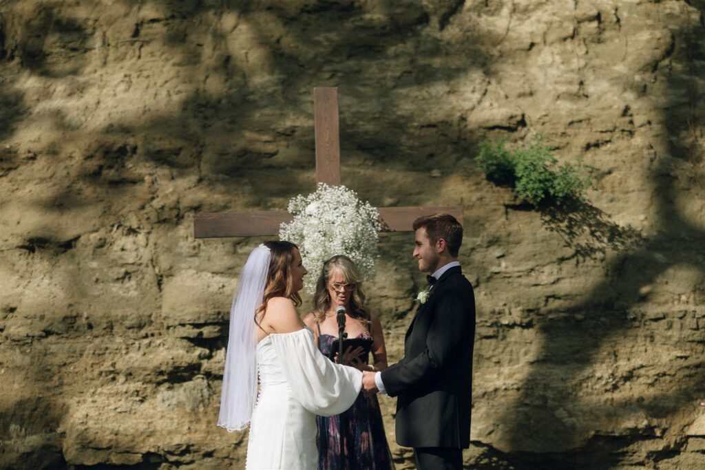 Bride and groom holding hands during their outdoor ceremony at Woods and Co Weddings and Events, standing beneath a wooden cross set against a stone backdrop.