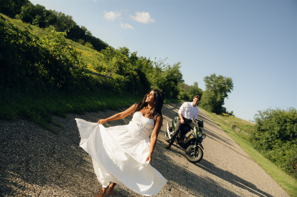 Amira leans back against a dark green moped on a quiet country road, barefoot in a white dress, looking up at Jake as he stands beside her in a white button-down during golden hour.