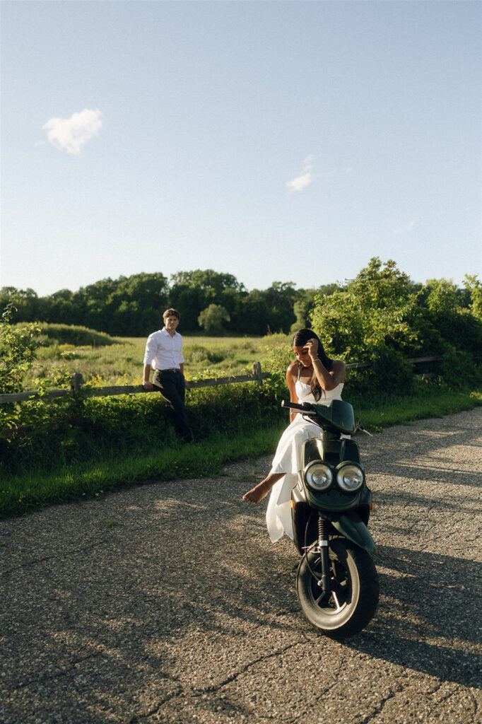 Amira sits sideways on the moped adjusting her hair while Jake leans against a wooden fence behind her, wide open Minnesota landscape stretching into the distance.