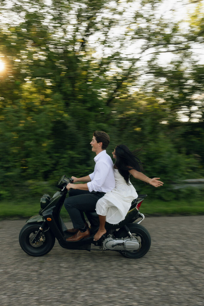 Amira throws her arms wide as Jake drives the moped down a tree-lined road at sunset, the motion blur adding a cinematic feel to their engagement photos.