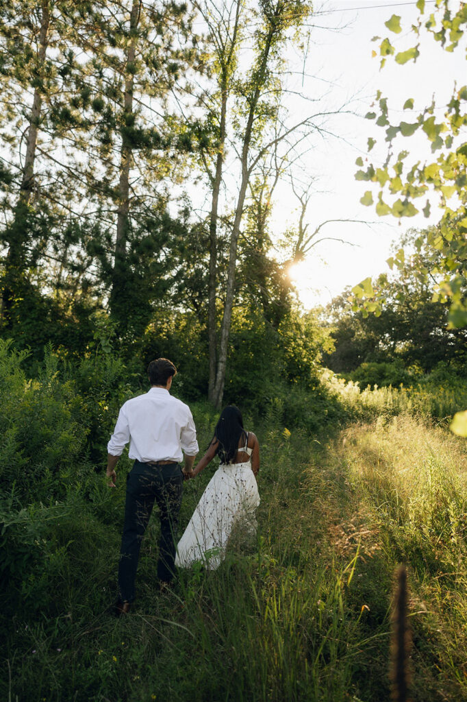 Amira and Jake walk hand in hand into tall green grass as golden hour light filters through the trees around them.