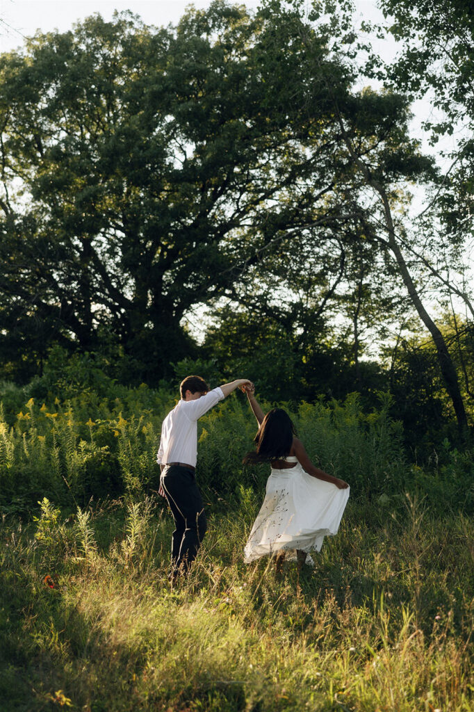 Jake spins Amira through a meadow at sunset, her white dress lifting slightly as warm light hits the grass around them.