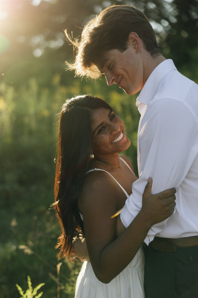 Close-up portrait of Amira smiling into the camera while Jake looks down at her, soft golden backlight catching her hair and shoulders.
