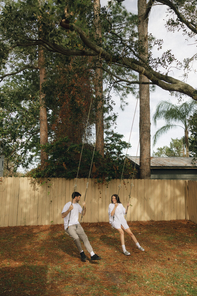 Couple swinging side by side in their backyard, sunlight filtering through tall trees as they laugh at each other mid-swing during their playful in-home Florida engagement session.