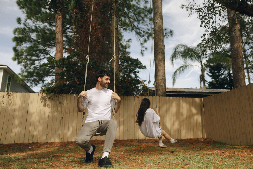 The two of them swinging out of sync on simple wooden swings, casual white outfits and bare yard creating that lived-in, cozy Florida engagement session vibe.