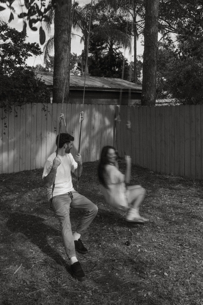 Black and white photo of her swinging forward in motion blur while he looks on, a cinematic moment from their relaxed in-home couples session in Florida.