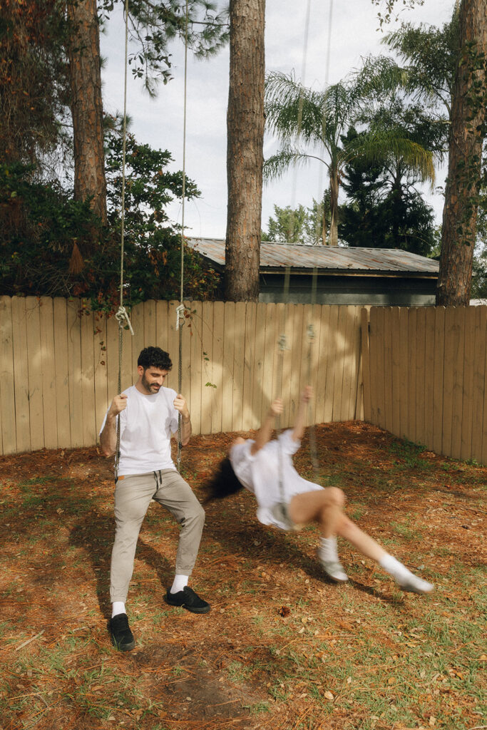 She swings back dramatically, legs extended and hair flying, while he steadies himself — playful movement captured during their backyard engagement session.