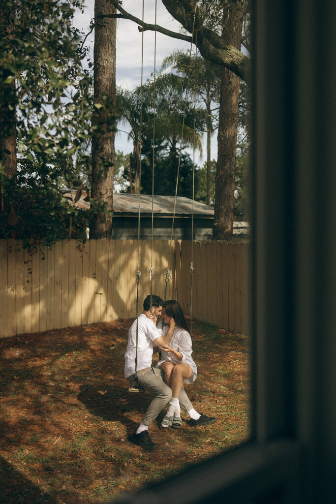 Framed through a window, the couple sits close together on the swings, foreheads touching in quiet intimacy during their cozy in-home Florida session.