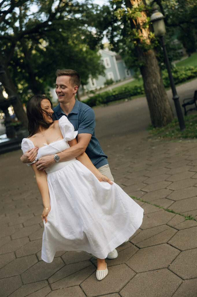 He wraps his arms around her from behind and spins her slightly on the paved walkway, both mid-laugh — playful, movement-heavy engagement photos in a quiet St. Paul neighborhood park.