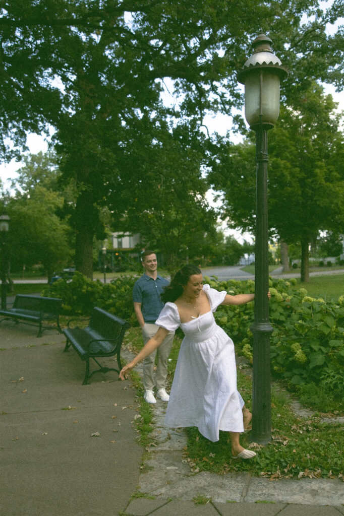 Playful engagement photo of a bride-to-be twirling around a lamppost while her fiancé watches, taken during a St. Paul park engagement session.