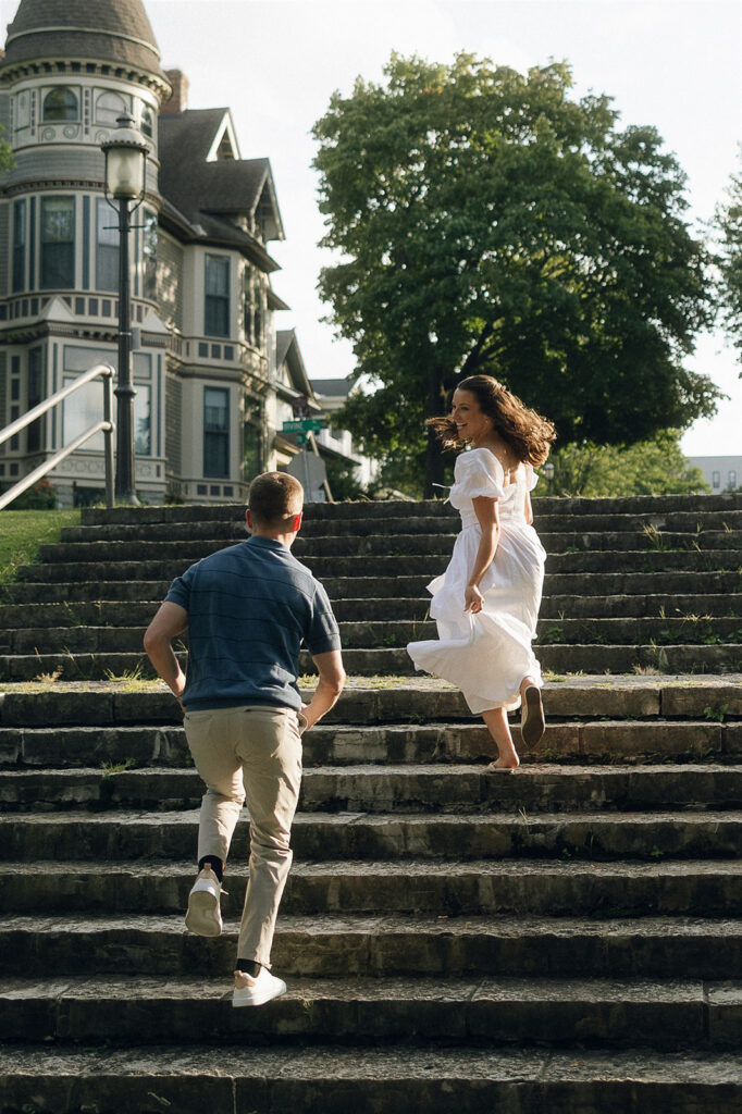 Couple running up the historic stone steps in St. Paul during their engagement session, her white dress catching the breeze as he chases after her in golden hour light.