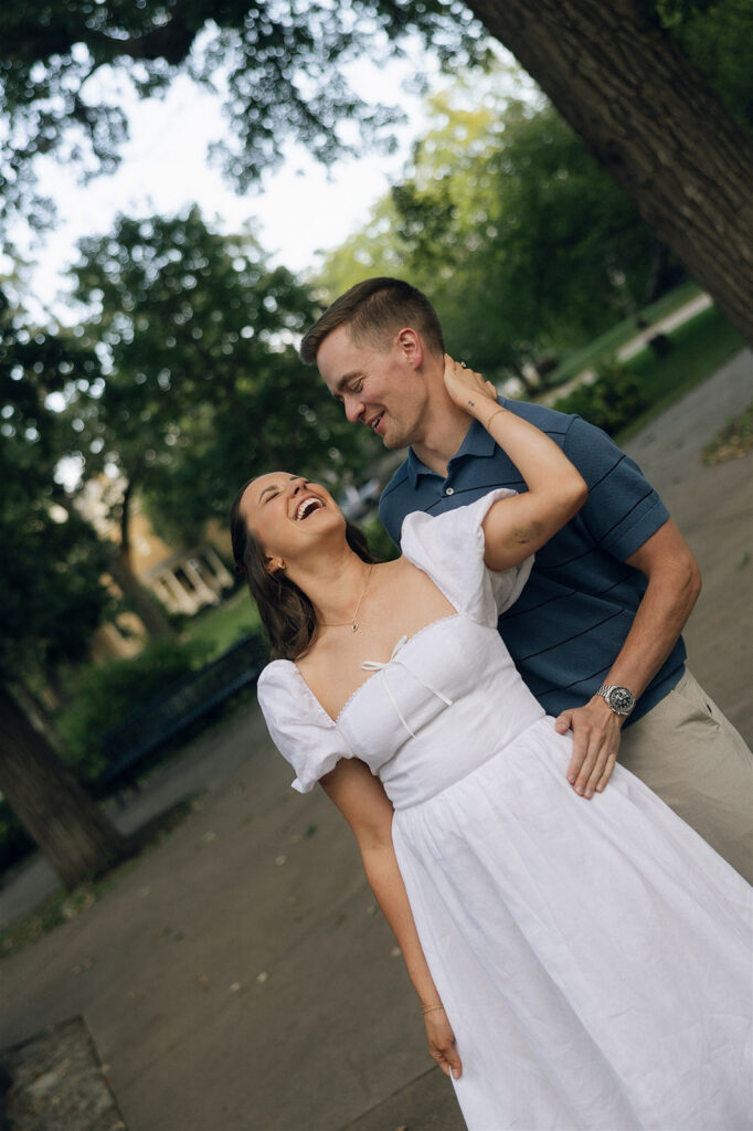 She throws her head back laughing as he pulls her in close along a tree-lined St. Paul park path, her white dress catching the movement while he looks at her like he already won.