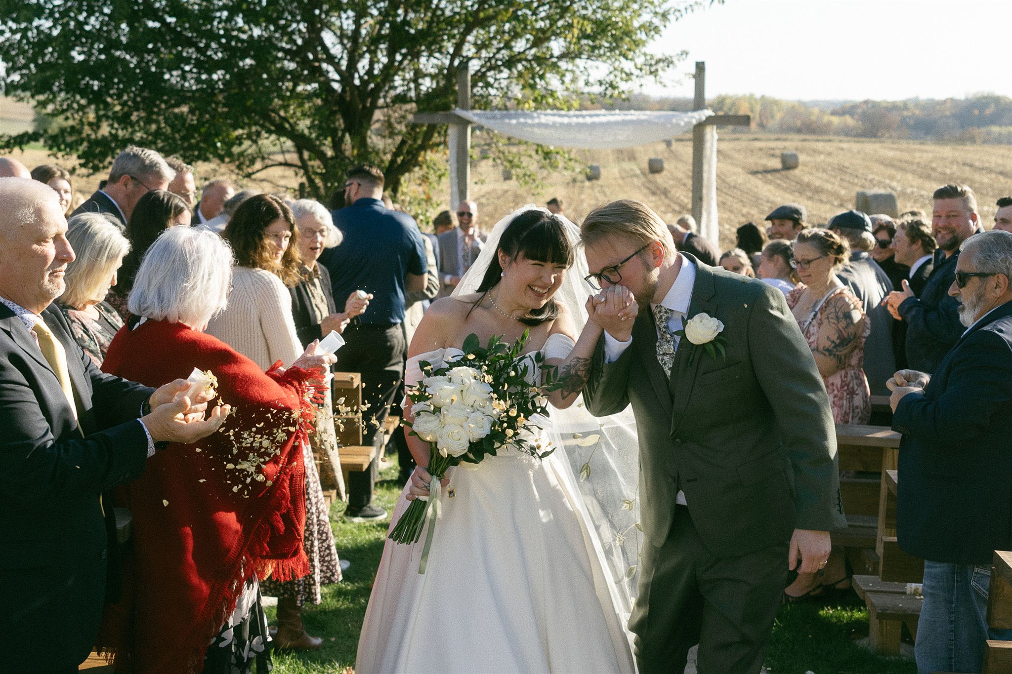 Newlyweds walking back down the aisle together at their outdoor Legacy Hill Farm ceremony, guests tossing petals and laughing around them.
