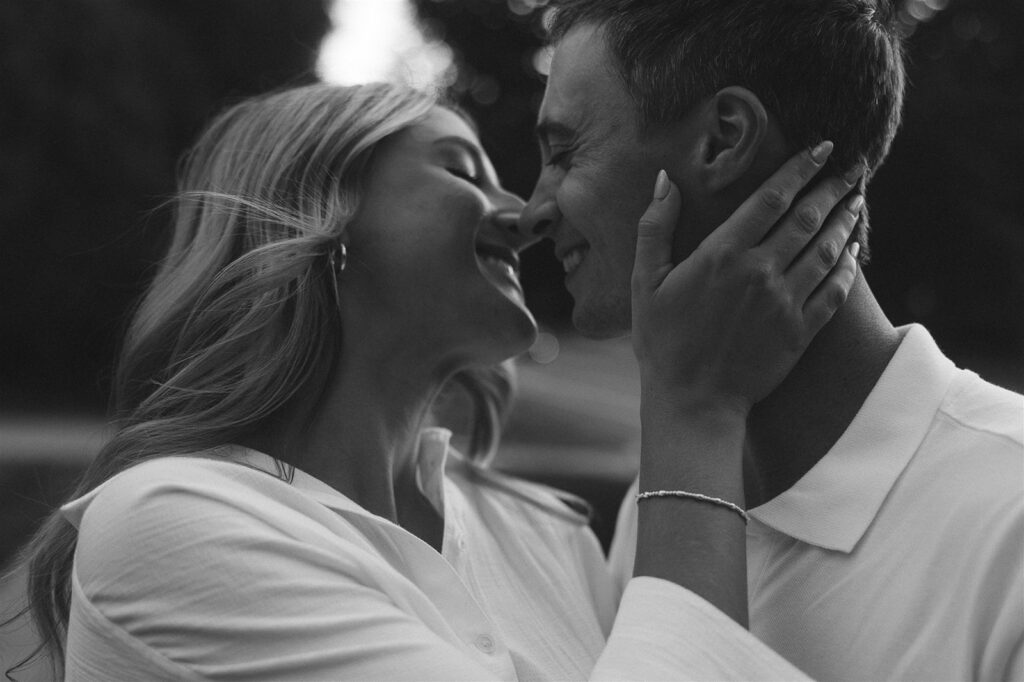 Close black and white image of them nearly nose-to-nose, her hand cupping his jaw as they smile right before a kiss.