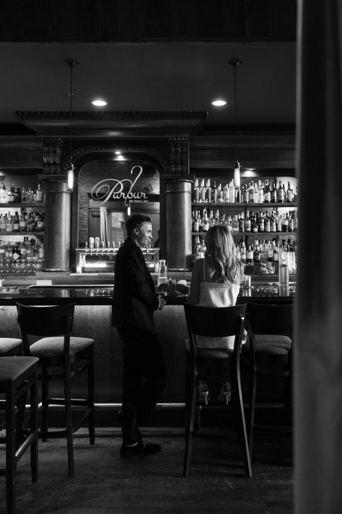 Black and white photo of an engaged couple sitting at the bar inside Parlor in St. Paul, sharing drinks and an easy laugh during their date-night-inspired engagement session.