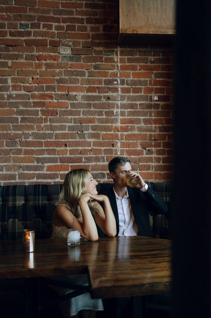 Engaged couple seated at a candlelit table in downtown St. Paul, she watches him sip his drink during their cozy indoor engagement session.