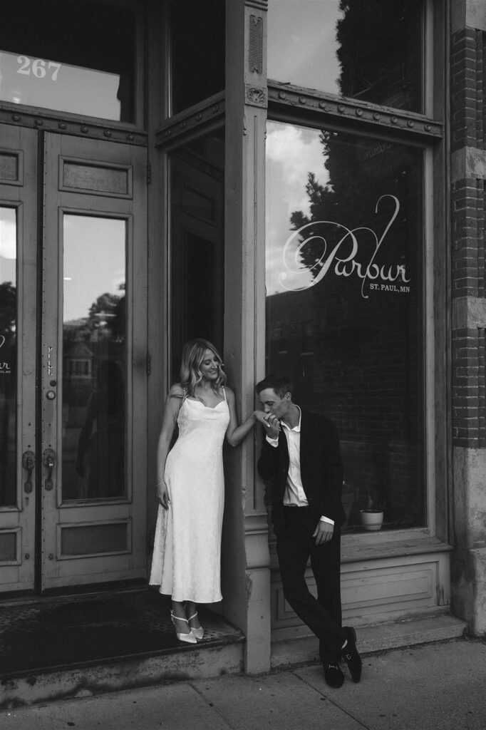 Black and white portrait of an engaged couple outside Parlor in St. Paul, he kisses her hand on the sidewalk during their city engagement session.