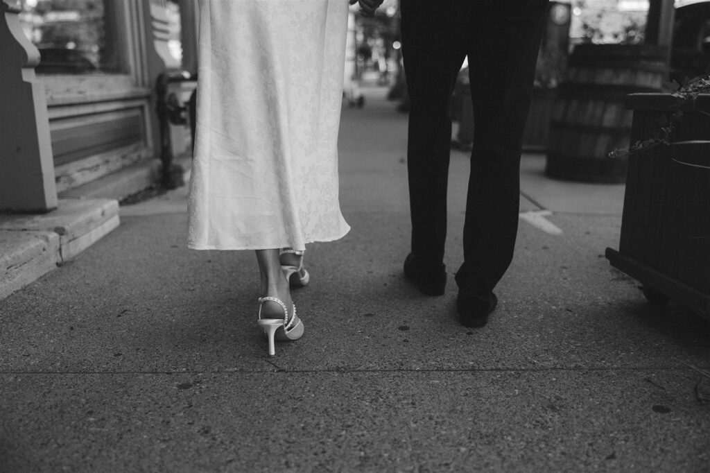 Black and white detail photo of a bride-to-be’s heels and dress hem walking beside her fiancé on a St. Paul sidewalk during their engagement session.